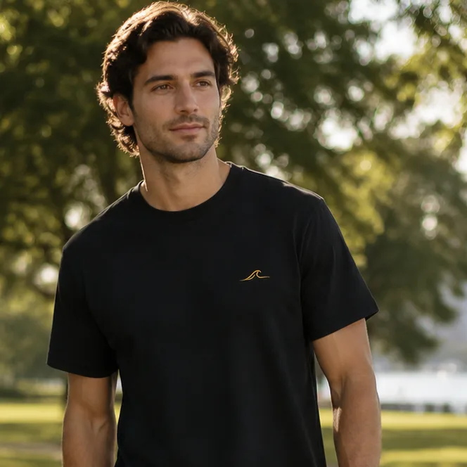 A man standing under the fig trees at Orleigh Park, West End, on the Brisbane River in the late afternoon, wearing a black 28° South tee with the gold wave mark embroidered on the chest.