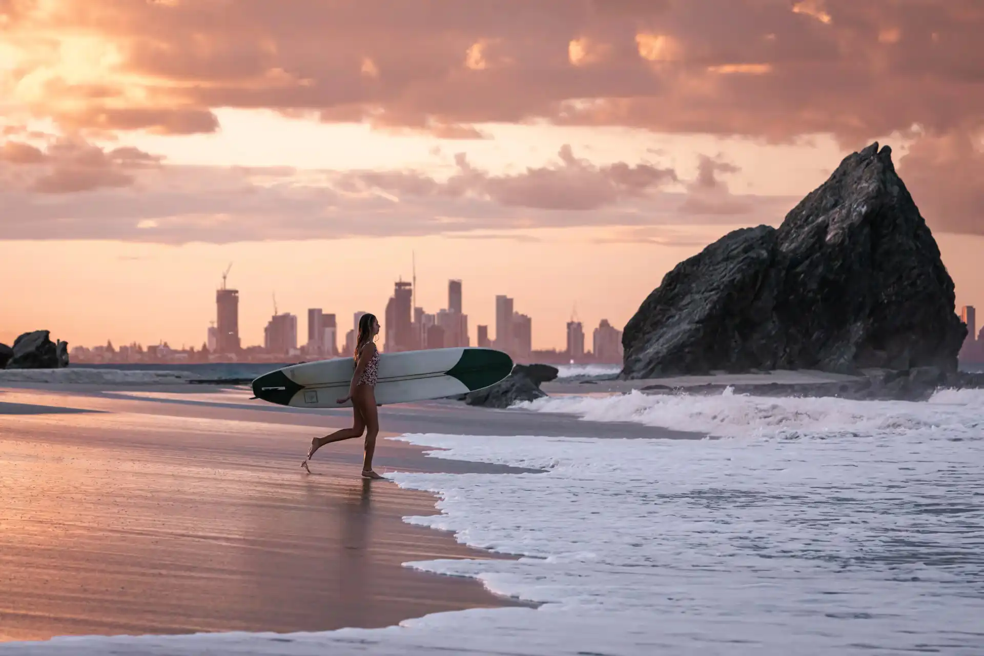 A female surfer carrying a surfboard past Elephant Rock in Currumbin at sunset, with the Gold Coast city skyline in the distant background.