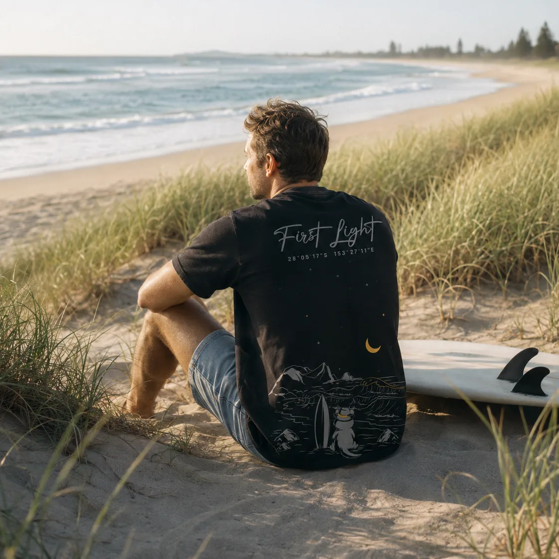 A man sitting in the dunes beside his surfboard looking out over an empty beach, wearing the 28° South First Light black tee with its moonlit shoreline back print.