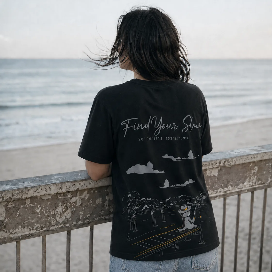 A woman leaning on a beachfront railing at Coolangatta, Gold Coast, wearing the 28° South Find Your Slow black tee. The back print shows Sunny the Quokka fishing off a jetty beneath the coordinates 28°06'15"S 153°27'09"E.